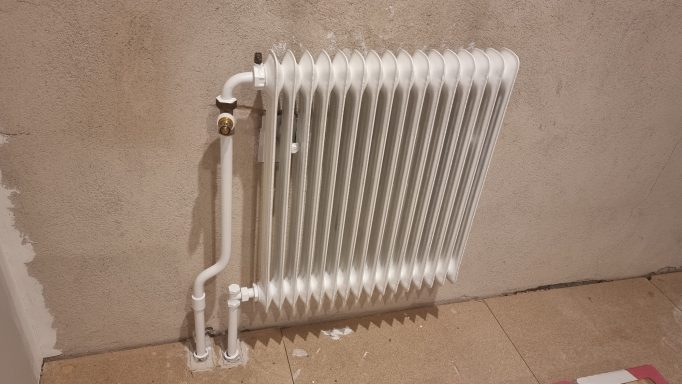 Vit radiator i gammaldags stil monterad på vägg, med rör anslutna.